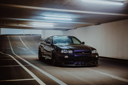 Der Nissan Skyline R34 steht tief und breit in der Kurve eines Parkhauses; Front, Seitenschweller und violette Akzente sind klar sichtbar. Originalmotiv „Violet Story“ im Querformat.