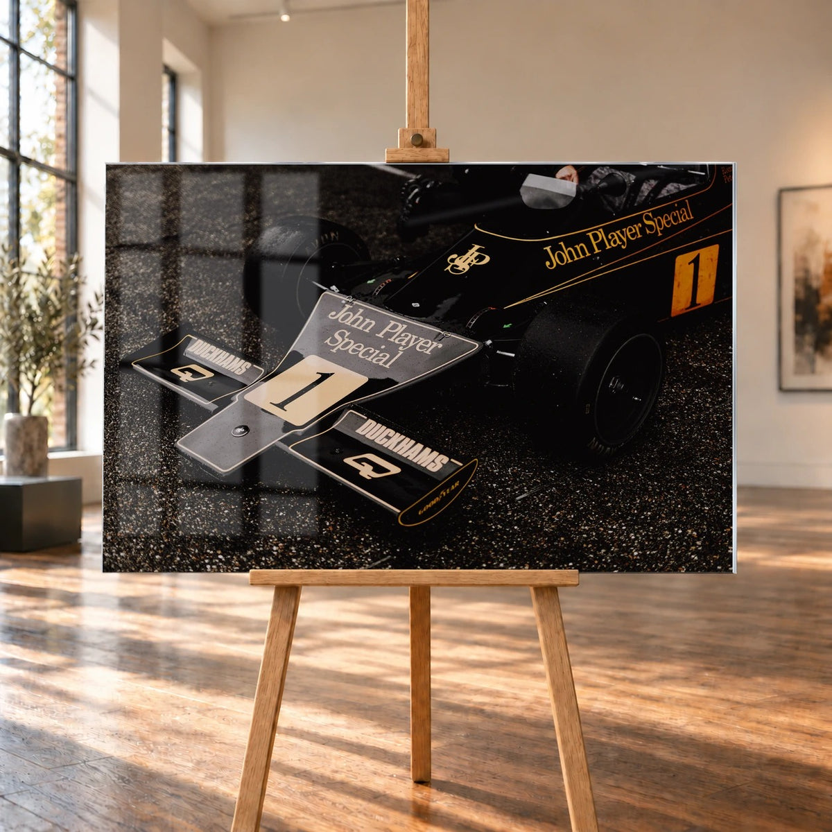 Die Nahaufnahme zeigt die spitze Front des Lotus 76 mit goldener John-Player-Special-Lackierung und großer Startnummer 1. Motiv „Apex Story“ als Acrylglas-Wandbild, auf einer Holzstaffelei in einem hellen Galerieraum.