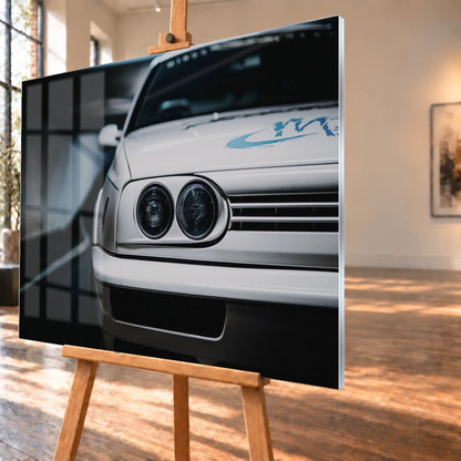 Die Frontpartie von Jesses Jetta ist eng angeschnitten; zwei runde Scheinwerfer und die graue Grafik auf weißem Lack bestimmen das Bild. Motiv „Silver Horizon“ als Acrylglas-Wandbild, schräg von der Seite auf einer Holzstaffelei.
