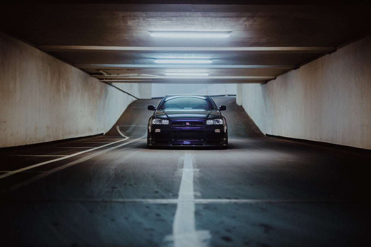 Der violette Nissan Skyline R34 steht frontal und klein am Ende einer langen, symmetrischen Parkhaustunnel-Achse; die helle Mittellinie führt direkt auf das Fahrzeug zu. Originalmotiv „Purple Depth“ im Querformat.