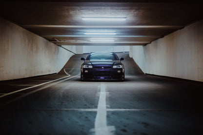 Der violette Nissan Skyline R34 steht frontal und klein am Ende einer langen, symmetrischen Parkhaustunnel-Achse; die helle Mittellinie führt direkt auf das Fahrzeug zu. Originalmotiv „Purple Depth“ im Querformat.