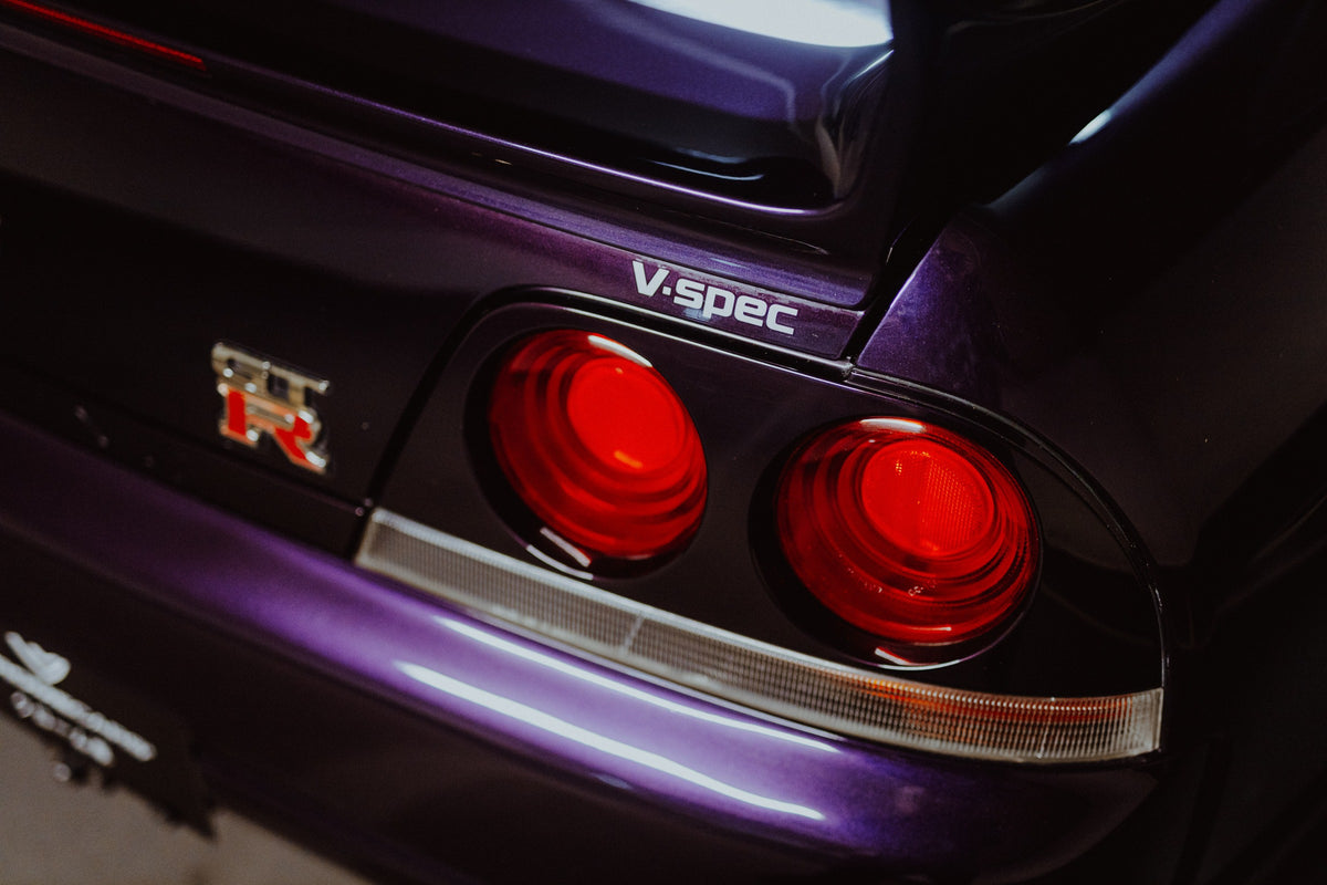 Eine enge Heckaufnahme zeigt zwei rote Rundlichter, GT-R- und V-Spec-Schriftzug sowie den violetten Lack des Nissan Skyline R33. Originalmotiv „Neon Presence“ im Querformat.