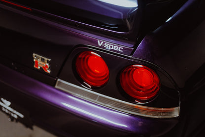 Eine enge Heckaufnahme zeigt zwei rote Rundlichter, GT-R- und V-Spec-Schriftzug sowie den violetten Lack des Nissan Skyline R33. Originalmotiv „Neon Presence“ im Querformat.