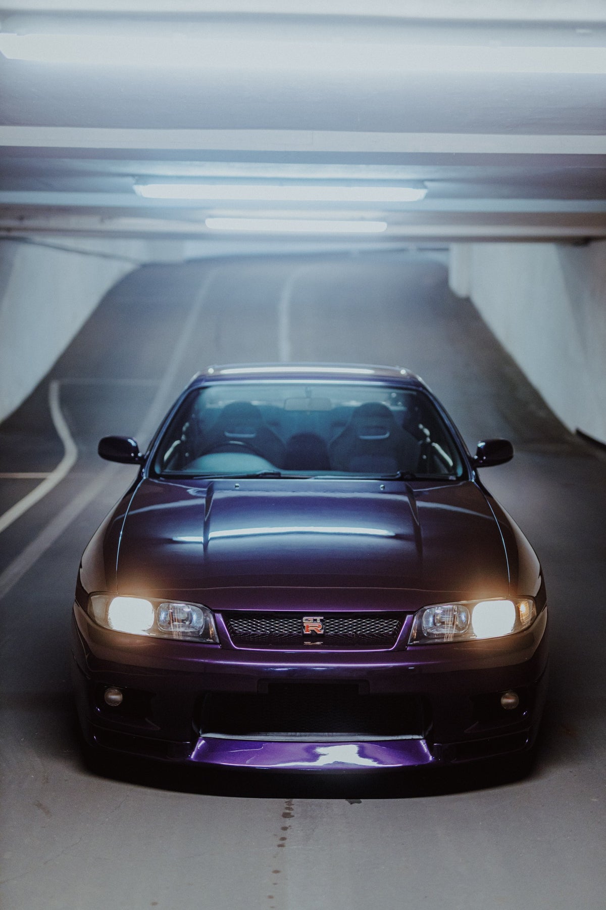 Die Front des Nissan Skyline R33 füllt das Hochformat; Scheinwerfer, Kühlergrill und der violette Frontsplitter liegen im kalten Deckenlicht. Originalmotiv „Neon Pulse“ im Hochformat.