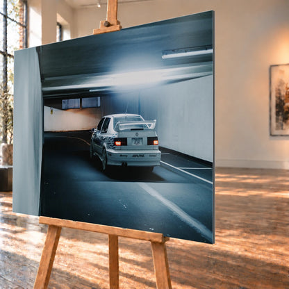 Jesses Jetta ist von hinten in einer langen Parkhausspur aufgenommen; die weiße Karosserie hebt sich klein vom kühlen Tunnel ab. Motiv „Screen Presence“ als Alu gebürstet-Wandbild, schräg von der Seite auf einer Holzstaffelei.