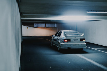 Jesses Jetta ist von hinten in einer langen Parkhausspur aufgenommen; die weiße Karosserie hebt sich klein vom kühlen Tunnel ab. Originalmotiv „Screen Presence“ im Querformat.