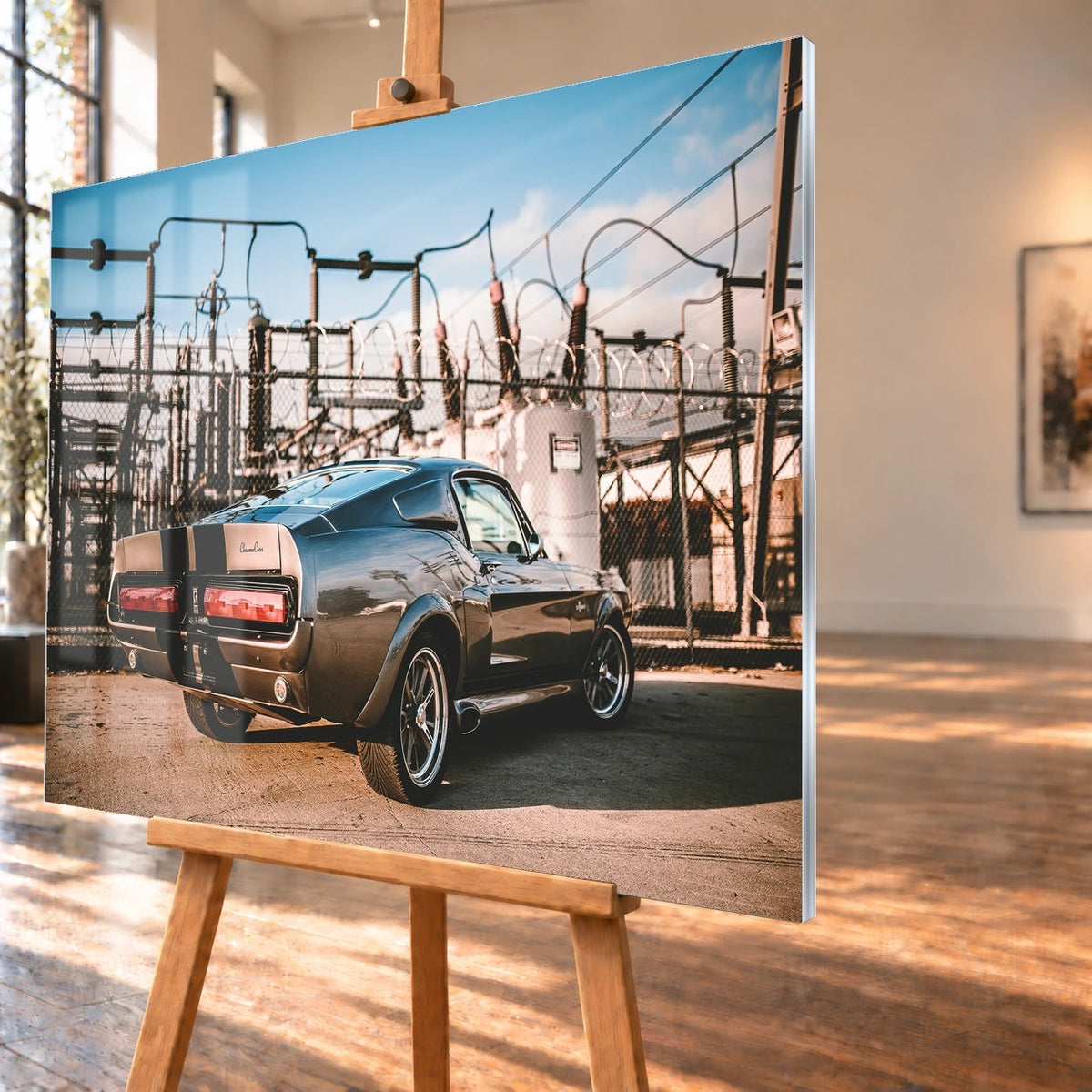 Eleanor steht schräg von hinten vor einem Umspannwerk; Masten, Leitungen und warme Sonne bilden einen technischen Hintergrund. Motiv „Road Moment“ als Acrylglas-Wandbild, schräg von der Seite auf einer Holzstaffelei.