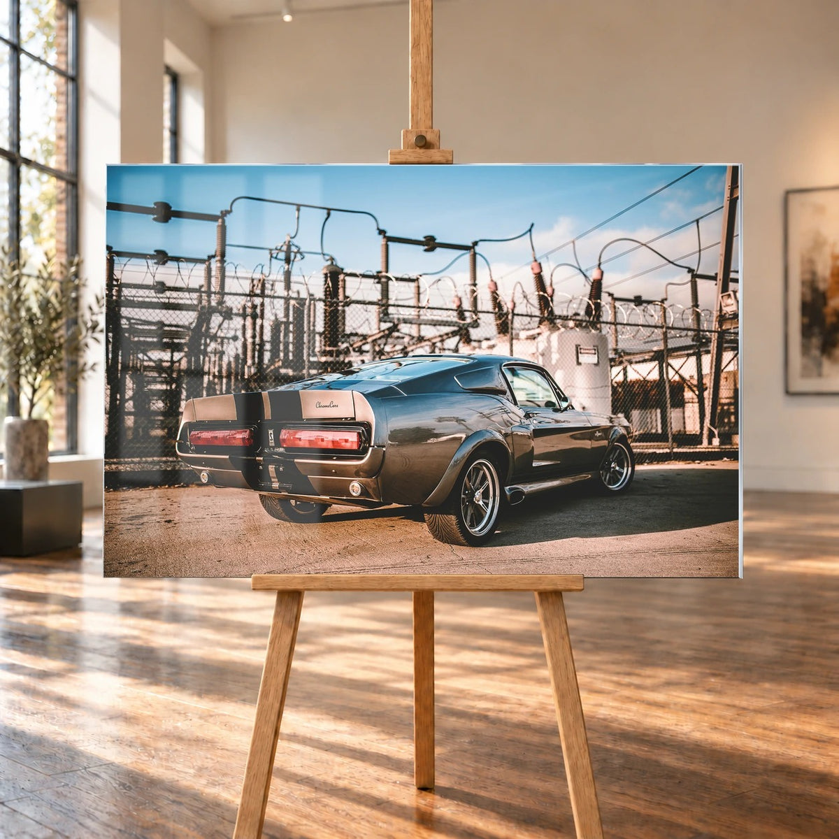 Eleanor steht schräg von hinten vor einem Umspannwerk; Masten, Leitungen und warme Sonne bilden einen technischen Hintergrund. Motiv „Road Moment“ als Acrylglas-Wandbild, auf einer Holzstaffelei in einem hellen Galerieraum.