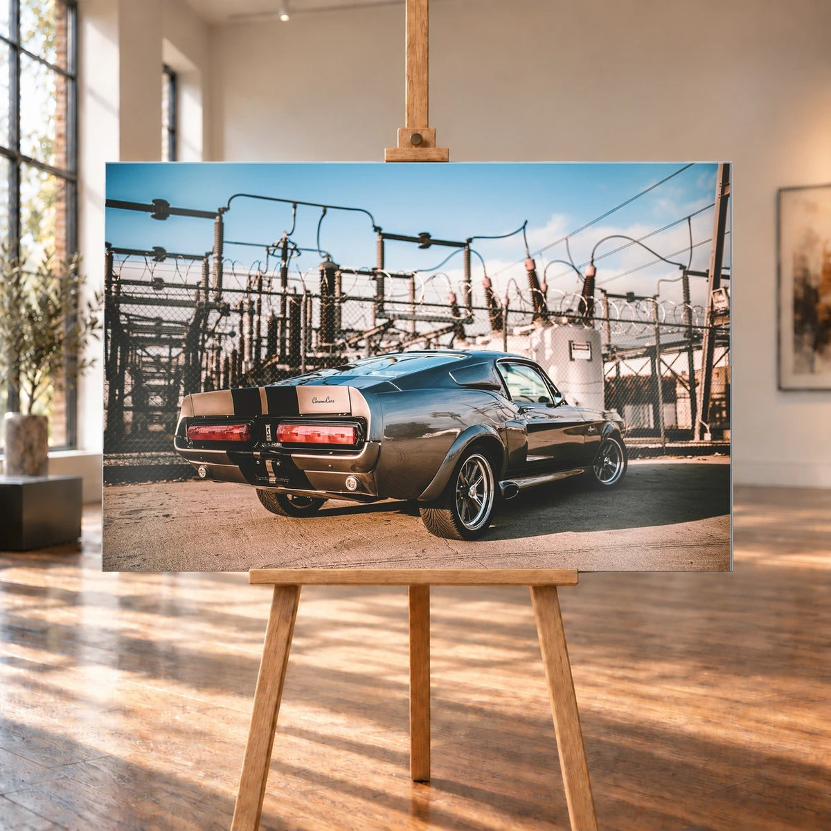 Eleanor steht schräg von hinten vor einem Umspannwerk; Masten, Leitungen und warme Sonne bilden einen technischen Hintergrund. Motiv „Road Moment“ als Alu gebürstet-Wandbild, auf einer Holzstaffelei in einem hellen Galerieraum.