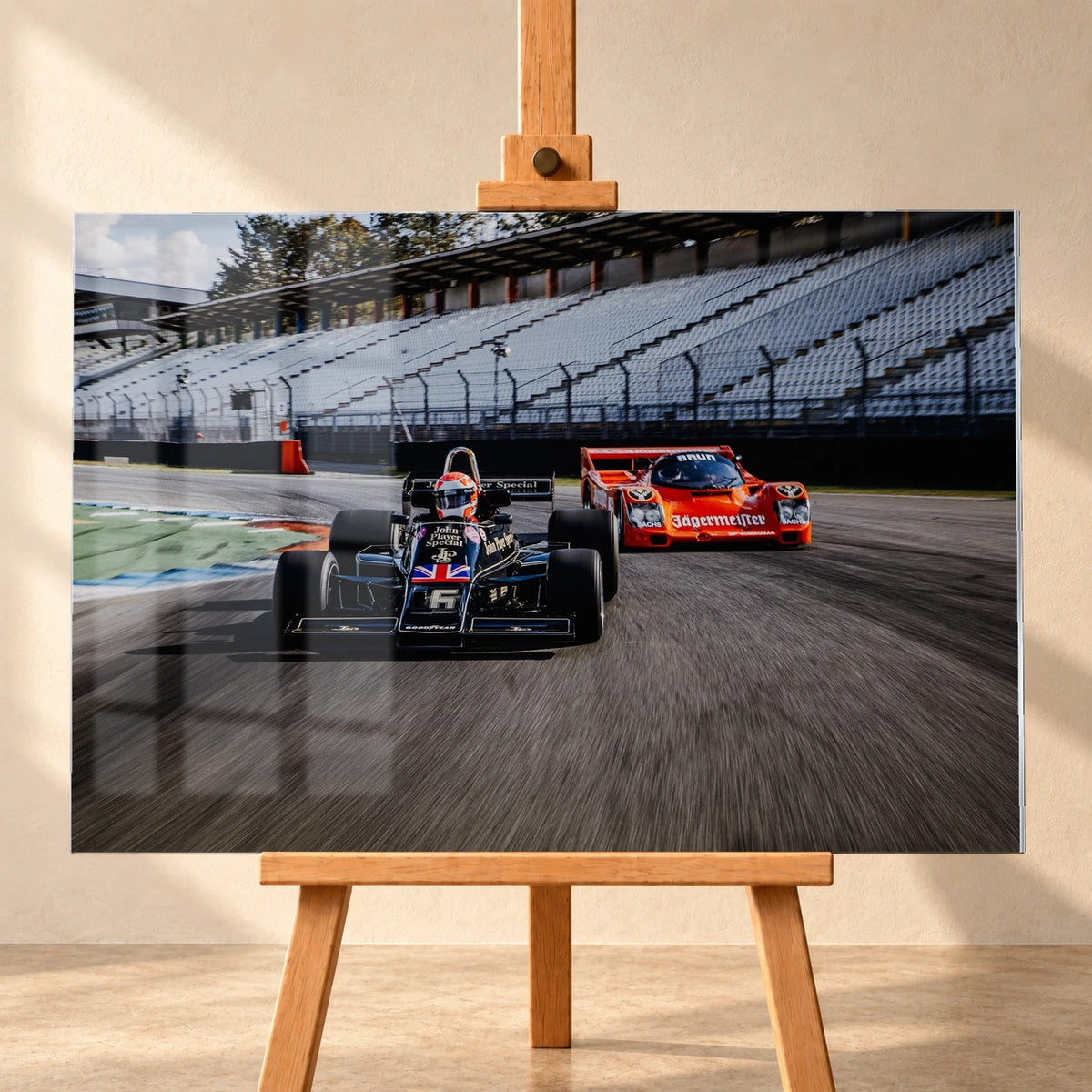 Lotus 77 und der Porsche 962 Brun/Jägermeister fahren nebeneinander beziehungsweise versetzt über die Strecke; orange und schwarz setzen klare Rennfarben. Motiv „Grid Rhythm“ als Acrylglas-Wandbild, frontal auf einer Holzstaffelei.