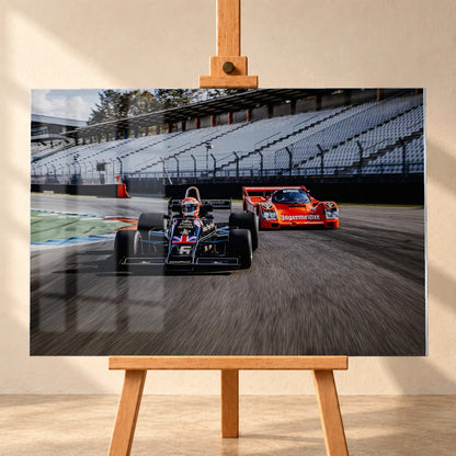 Lotus 77 und der Porsche 962 Brun/Jägermeister fahren nebeneinander beziehungsweise versetzt über die Strecke; orange und schwarz setzen klare Rennfarben. Motiv „Grid Rhythm“ als Acrylglas-Wandbild, frontal auf einer Holzstaffelei.