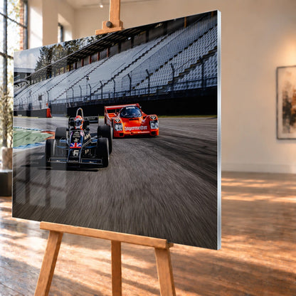 Lotus 77 und der Porsche 962 Brun/Jägermeister fahren nebeneinander beziehungsweise versetzt über die Strecke; orange und schwarz setzen klare Rennfarben. Motiv „Grid Rhythm“ als Acrylglas-Wandbild, schräg von der Seite auf einer Holzstaffelei.