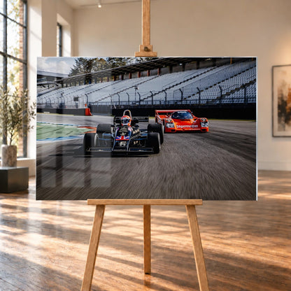 Lotus 77 und der Porsche 962 Brun/Jägermeister fahren nebeneinander beziehungsweise versetzt über die Strecke; orange und schwarz setzen klare Rennfarben. Motiv „Grid Rhythm“ als Acrylglas-Wandbild, auf einer Holzstaffelei in einem hellen Galerieraum.