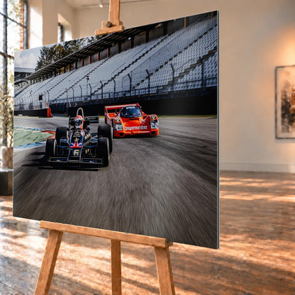 Lotus 77 und der Porsche 962 Brun/Jägermeister fahren nebeneinander beziehungsweise versetzt über die Strecke; orange und schwarz setzen klare Rennfarben. Motiv „Grid Rhythm“ als Alu gebürstet-Wandbild, schräg von der Seite auf einer Holzstaffelei.