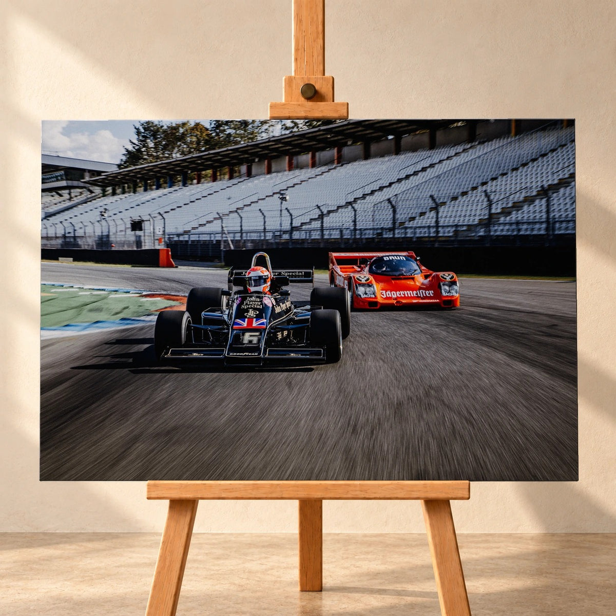 Lotus 77 und der Porsche 962 Brun/Jägermeister fahren nebeneinander beziehungsweise versetzt über die Strecke; orange und schwarz setzen klare Rennfarben. Motiv „Grid Rhythm“ als Leinwand-Wandbild, frontal auf einer Holzstaffelei.