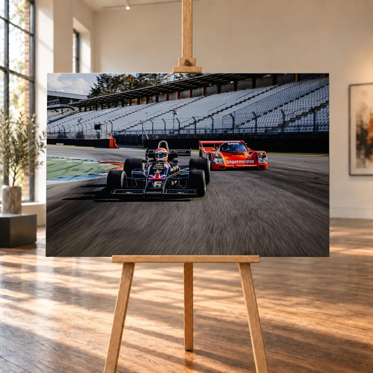 Lotus 77 und der Porsche 962 Brun/Jägermeister fahren nebeneinander beziehungsweise versetzt über die Strecke; orange und schwarz setzen klare Rennfarben. Motiv „Grid Rhythm“ als Leinwand-Wandbild, auf einer Holzstaffelei in einem hellen Galerieraum.