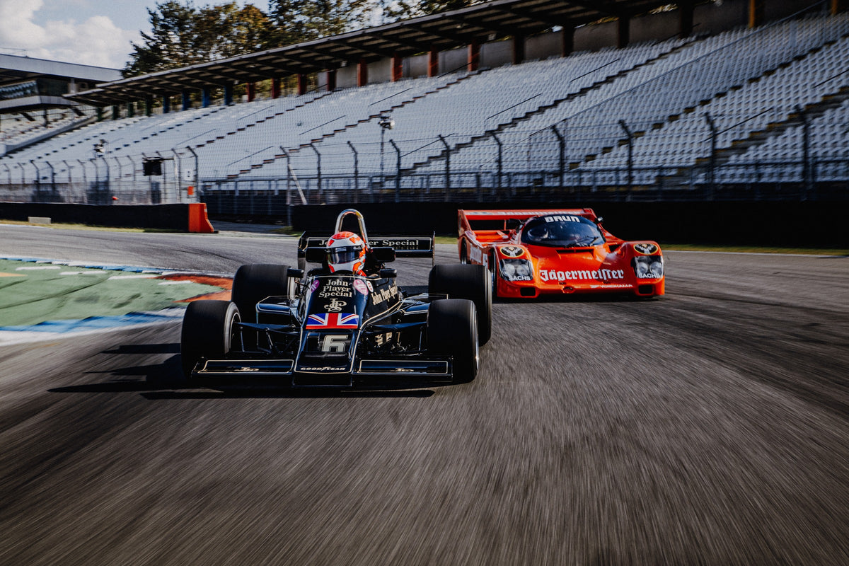 Lotus 77 und der Porsche 962 Brun/Jägermeister fahren nebeneinander beziehungsweise versetzt über die Strecke; orange und schwarz setzen klare Rennfarben. Originalmotiv „Grid Rhythm“ im Querformat.
