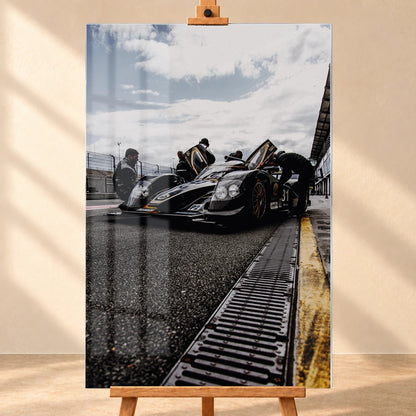 Ein Lotus LMP2 steht im Hochformat an der Boxenmauer, während Personen daneben arbeiten; Himmel und Pitlane öffnen die Szene nach oben. Motiv „Trackside Light“ als Acrylglas-Wandbild, frontal auf einer Holzstaffelei.