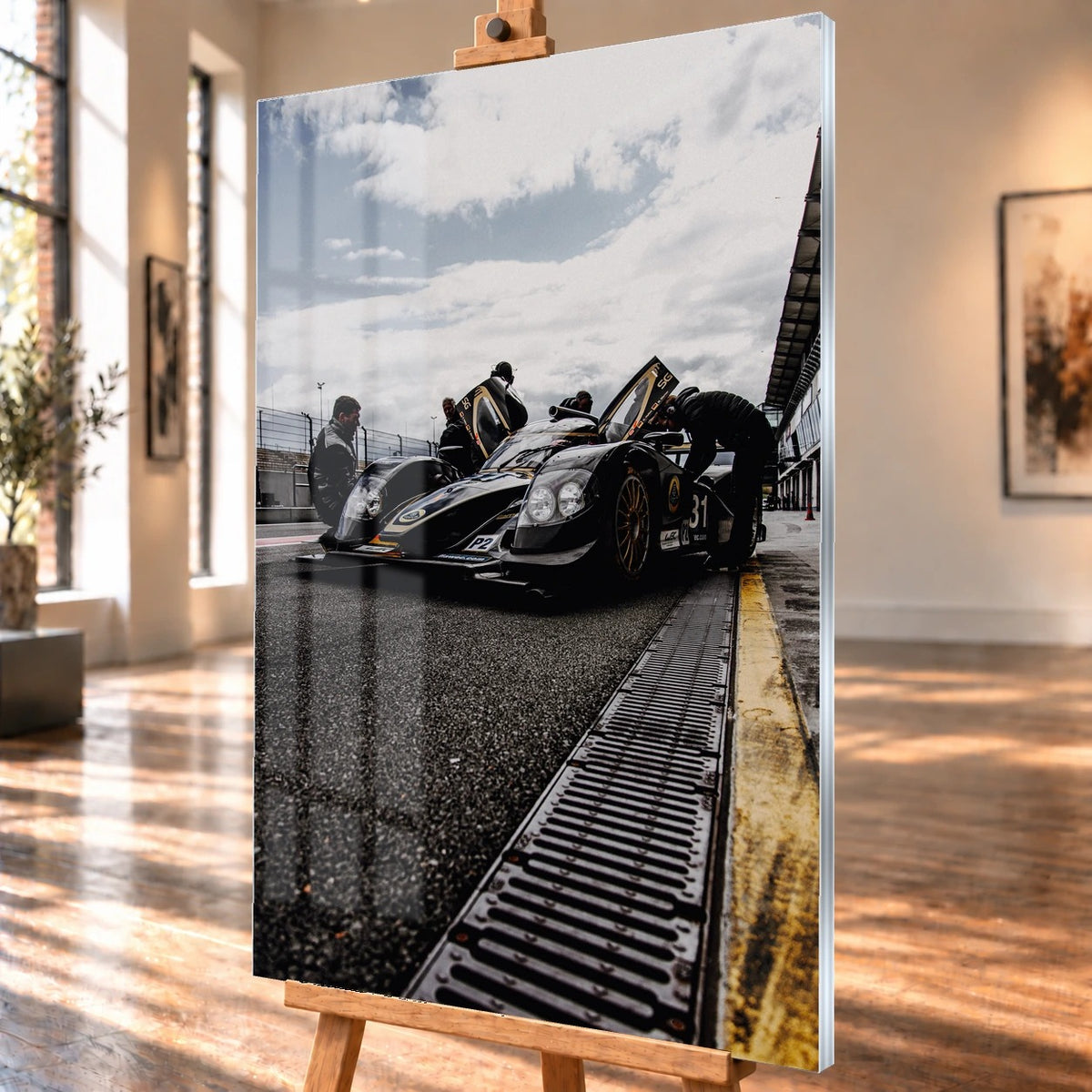 Ein Lotus LMP2 steht im Hochformat an der Boxenmauer, während Personen daneben arbeiten; Himmel und Pitlane öffnen die Szene nach oben. Motiv „Trackside Light“ als Acrylglas-Wandbild, schräg von der Seite auf einer Holzstaffelei.