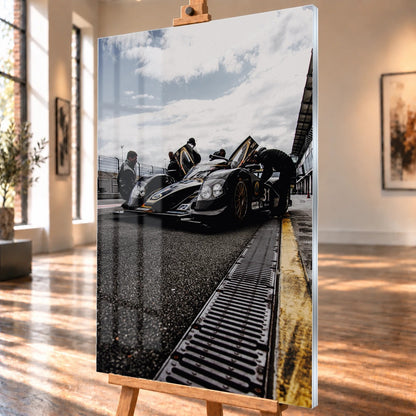 Ein Lotus LMP2 steht im Hochformat an der Boxenmauer, während Personen daneben arbeiten; Himmel und Pitlane öffnen die Szene nach oben. Motiv „Trackside Light“ als Acrylglas-Wandbild, schräg von der Seite auf einer Holzstaffelei.