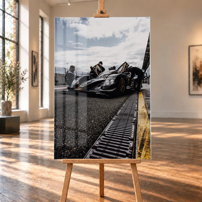 Ein Lotus LMP2 steht im Hochformat an der Boxenmauer, während Personen daneben arbeiten; Himmel und Pitlane öffnen die Szene nach oben. Motiv „Trackside Light“ als Acrylglas-Wandbild, auf einer Holzstaffelei in einem hellen Galerieraum.