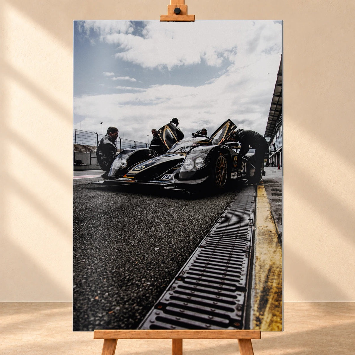 Ein Lotus LMP2 steht im Hochformat an der Boxenmauer, während Personen daneben arbeiten; Himmel und Pitlane öffnen die Szene nach oben. Motiv „Trackside Light“ als Alu gebürstet-Wandbild, frontal auf einer Holzstaffelei.