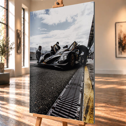 Ein Lotus LMP2 steht im Hochformat an der Boxenmauer, während Personen daneben arbeiten; Himmel und Pitlane öffnen die Szene nach oben. Motiv „Trackside Light“ als Alu gebürstet-Wandbild, schräg von der Seite auf einer Holzstaffelei.