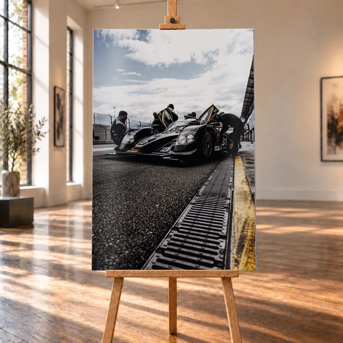 Ein Lotus LMP2 steht im Hochformat an der Boxenmauer, während Personen daneben arbeiten; Himmel und Pitlane öffnen die Szene nach oben. Motiv „Trackside Light“ als Alu gebürstet-Wandbild, auf einer Holzstaffelei in einem hellen Galerieraum.