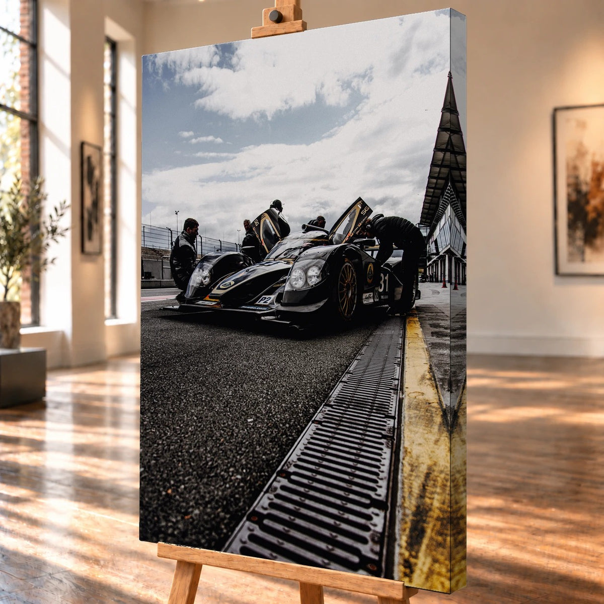 Ein Lotus LMP2 steht im Hochformat an der Boxenmauer, während Personen daneben arbeiten; Himmel und Pitlane öffnen die Szene nach oben. Motiv „Trackside Light“ als Leinwand-Wandbild, schräg von der Seite auf einer Holzstaffelei.