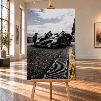Ein Lotus LMP2 steht im Hochformat an der Boxenmauer, während Personen daneben arbeiten; Himmel und Pitlane öffnen die Szene nach oben. Motiv „Trackside Light“ als Leinwand-Wandbild, auf einer Holzstaffelei in einem hellen Galerieraum.