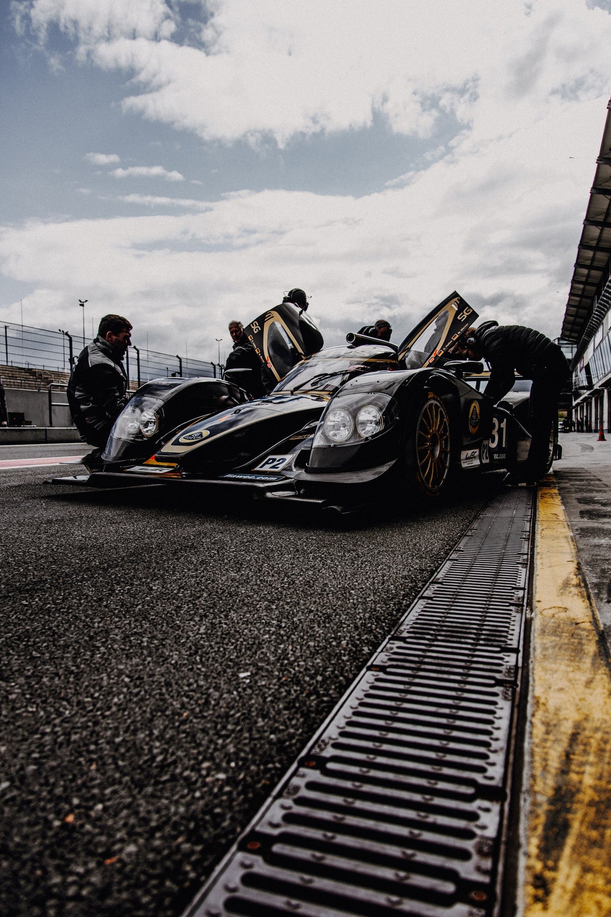 Ein Lotus LMP2 steht im Hochformat an der Boxenmauer, während Personen daneben arbeiten; Himmel und Pitlane öffnen die Szene nach oben. Originalmotiv „Trackside Light“ im Hochformat.