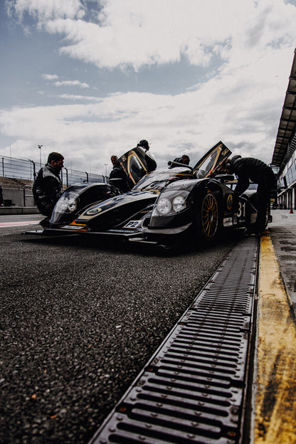 Ein Lotus LMP2 steht im Hochformat an der Boxenmauer, während Personen daneben arbeiten; Himmel und Pitlane öffnen die Szene nach oben. Originalmotiv „Trackside Light“ im Hochformat.