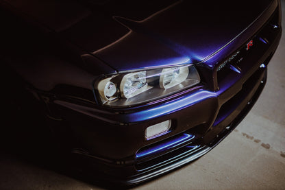 Eine schräge Nahaufnahme zeigt Scheinwerfer, Frontschürze und violette Lackkante des Nissan Skyline R34 unter hartem Neonlicht. Originalmotiv „Skyline Presence“ im Querformat.