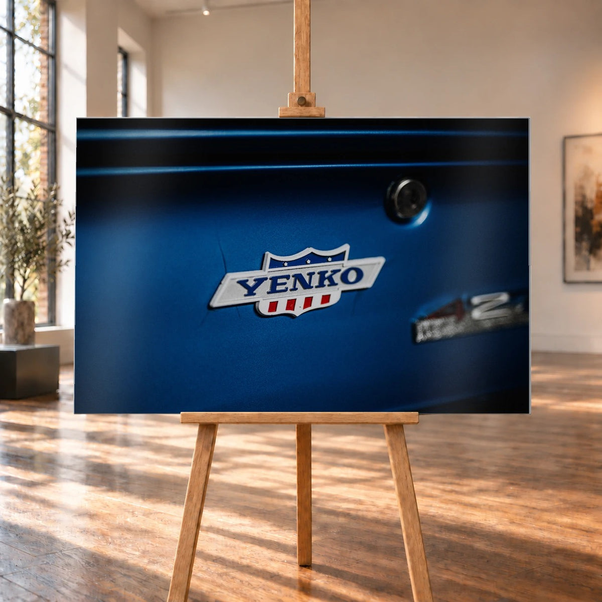 Das Yenko-Emblem sitzt als helles Detail auf der blauen Karosserie; Lack, Chrom und Schriftzug füllen die Aufnahme. Motiv „Hero Line“ als Alu gebürstet-Wandbild, auf einer Holzstaffelei in einem hellen Galerieraum.