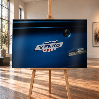Das Yenko-Emblem sitzt als helles Detail auf der blauen Karosserie; Lack, Chrom und Schriftzug füllen die Aufnahme. Motiv „Hero Line“ als Alu gebürstet-Wandbild, auf einer Holzstaffelei in einem hellen Galerieraum.