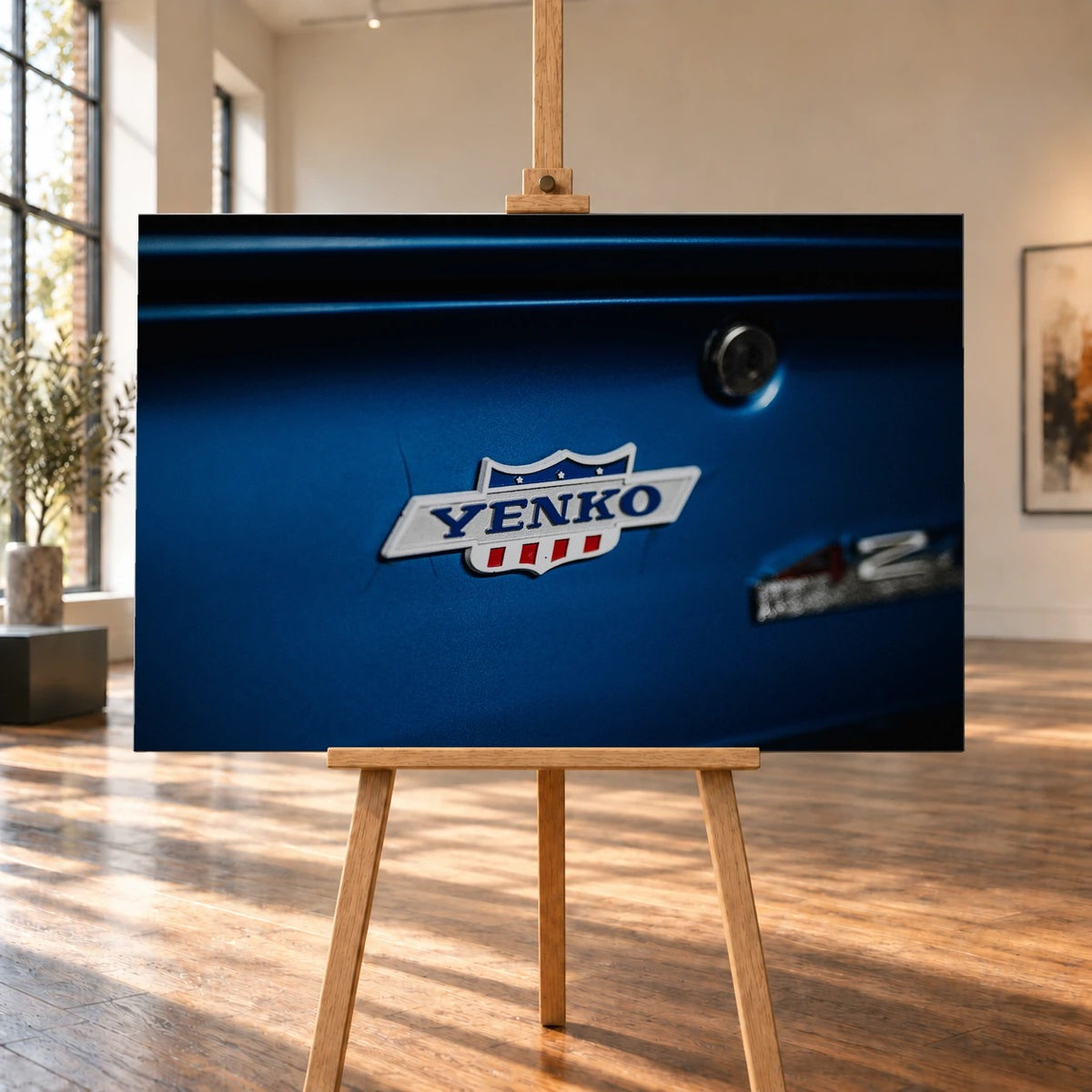 Das Yenko-Emblem sitzt als helles Detail auf der blauen Karosserie; Lack, Chrom und Schriftzug füllen die Aufnahme. Motiv „Hero Line“ als Leinwand-Wandbild, auf einer Holzstaffelei in einem hellen Galerieraum.