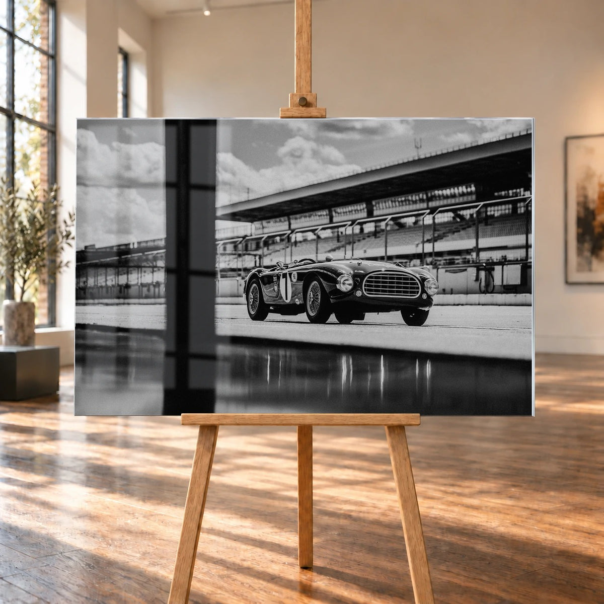 Der MG Tojeiro steht im Schwarz-Weiß-Seitenprofil vor der Boxenanlage; sein Spiegelbild erscheint auf dem nassen Boden. Motiv „Trackside Echo“ als Acrylglas-Wandbild, auf einer Holzstaffelei in einem hellen Galerieraum.