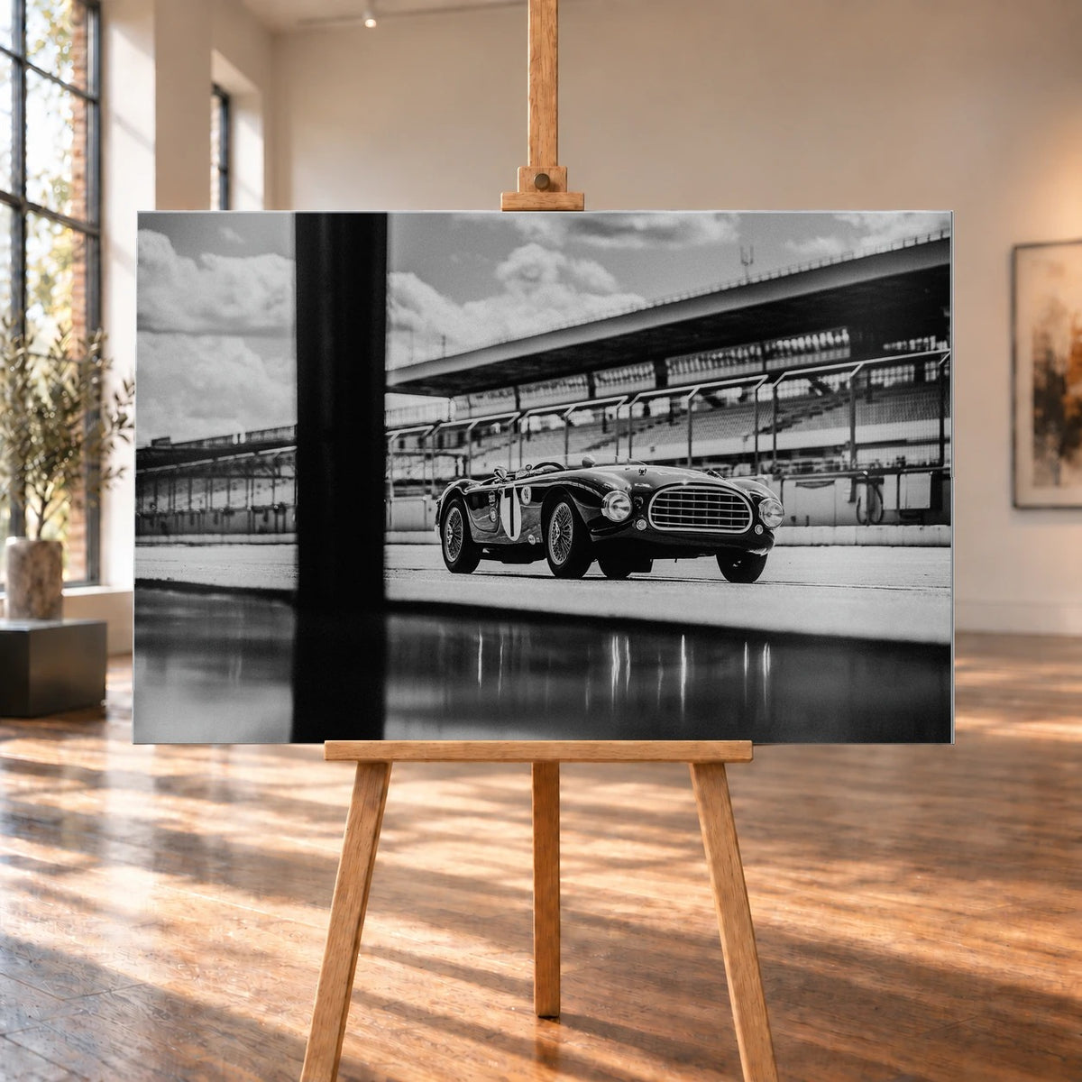 Der MG Tojeiro steht im Schwarz-Weiß-Seitenprofil vor der Boxenanlage; sein Spiegelbild erscheint auf dem nassen Boden. Motiv „Trackside Echo“ als Alu gebürstet-Wandbild, auf einer Holzstaffelei in einem hellen Galerieraum.