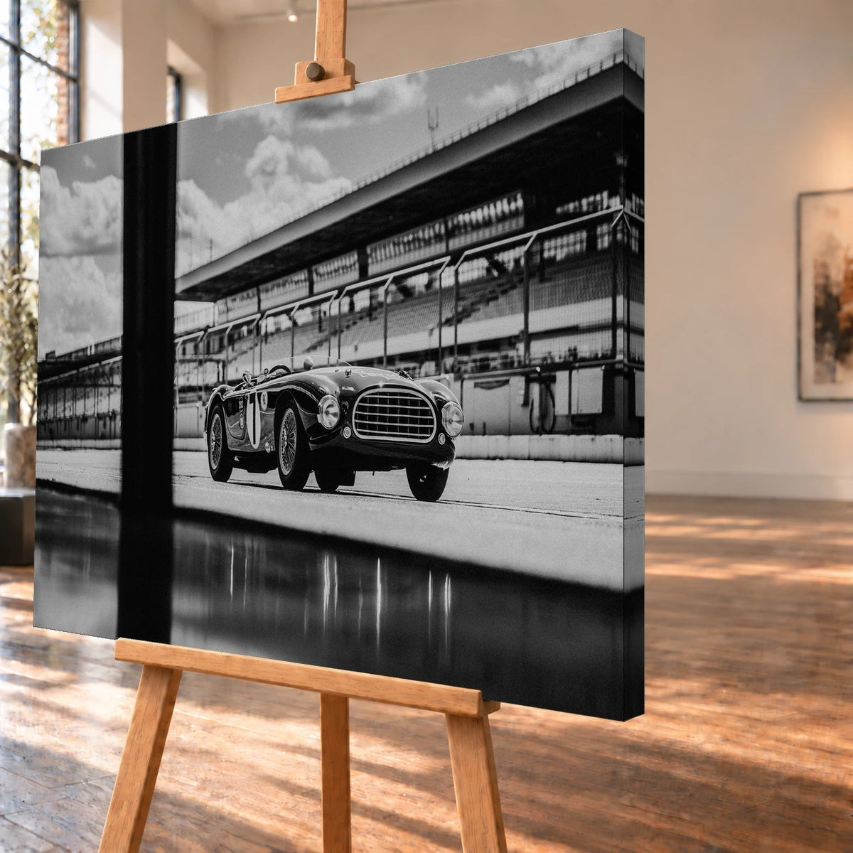 Der MG Tojeiro steht im Schwarz-Weiß-Seitenprofil vor der Boxenanlage; sein Spiegelbild erscheint auf dem nassen Boden. Motiv „Trackside Echo“ als Leinwand-Wandbild, schräg von der Seite auf einer Holzstaffelei.