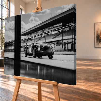 Der MG Tojeiro steht im Schwarz-Weiß-Seitenprofil vor der Boxenanlage; sein Spiegelbild erscheint auf dem nassen Boden. Motiv „Trackside Echo“ als Leinwand-Wandbild, schräg von der Seite auf einer Holzstaffelei.