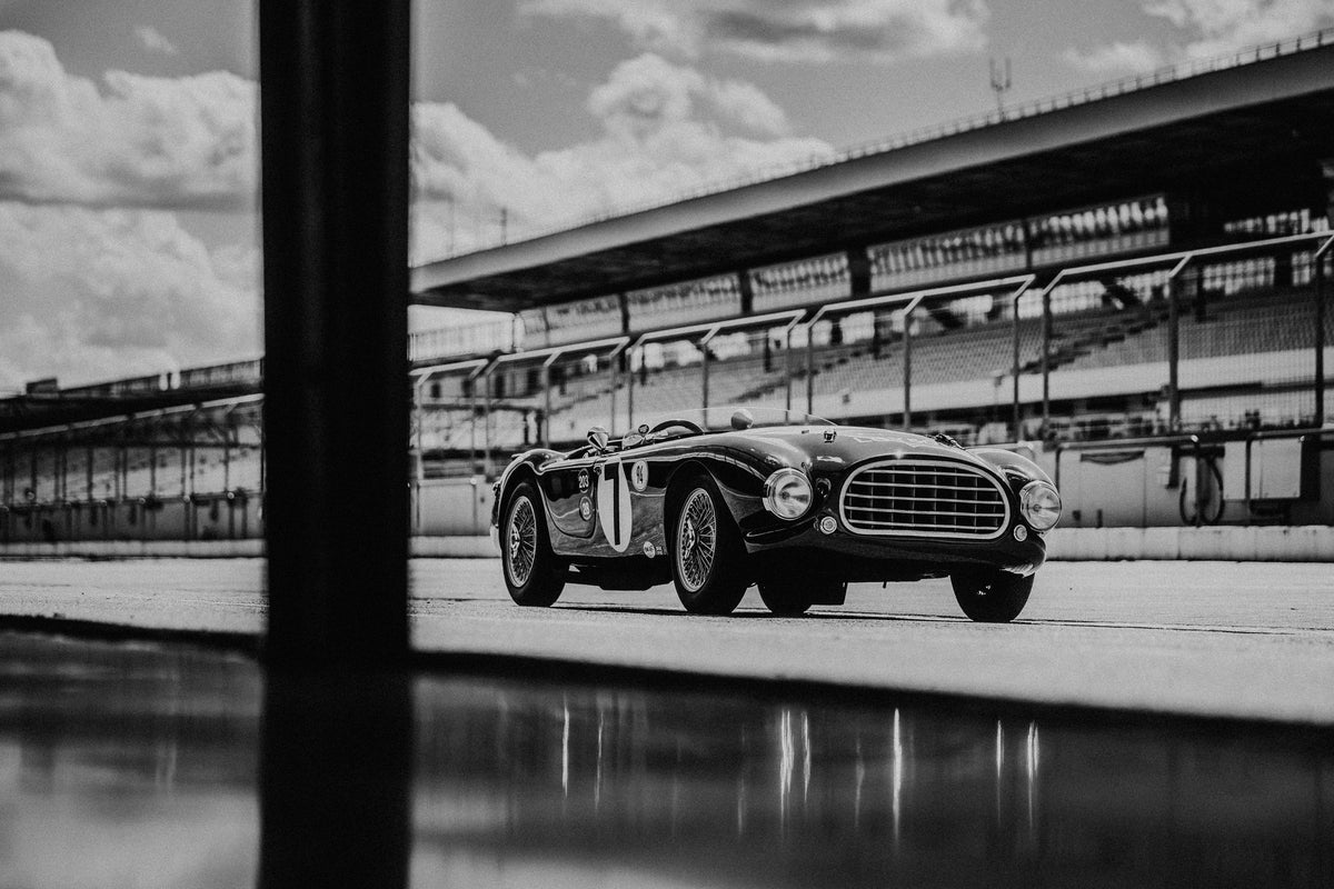 Der MG Tojeiro steht im Schwarz-Weiß-Seitenprofil vor der Boxenanlage; sein Spiegelbild erscheint auf dem nassen Boden. Originalmotiv „Trackside Echo“ im Querformat.