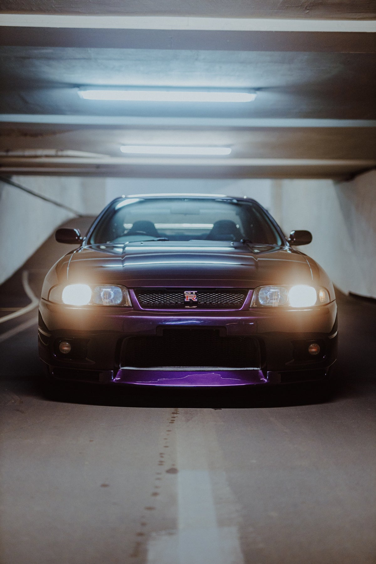 Die Front des Nissan Skyline R33 erscheint im Hochformat mit eingeschalteten Scheinwerfern; violetter Splitter und kaltes Neonlicht kontrastieren miteinander. Originalmotiv „Midnight Depth“ im Hochformat.