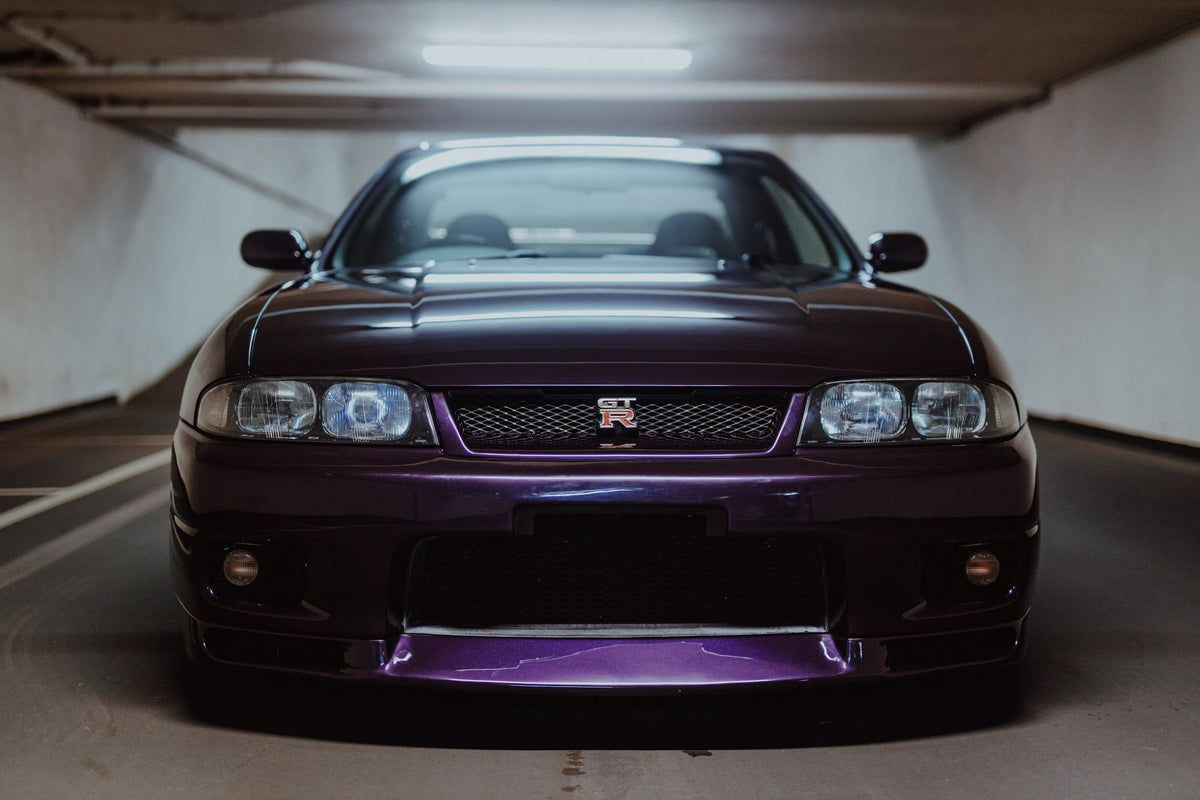 Der Nissan Skyline R33 ist frontal und nah unter Neonlicht fotografiert; Kühlergrill und violetter Splitter bilden eine klare horizontale Linie. Originalmotiv „Skyline Moment“ im Querformat.