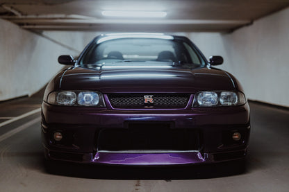 Der Nissan Skyline R33 ist frontal und nah unter Neonlicht fotografiert; Kühlergrill und violetter Splitter bilden eine klare horizontale Linie. Originalmotiv „Skyline Moment“ im Querformat.