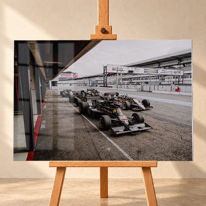 Mehrere historische Lotus-Rennwagen rollen in enger Formation an der Boxengasse entlang; der Blick von oben zeigt die Staffelung. Motiv „Trackside Moment“ als Acrylglas-Wandbild, frontal auf einer Holzstaffelei.