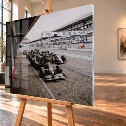 Mehrere historische Lotus-Rennwagen rollen in enger Formation an der Boxengasse entlang; der Blick von oben zeigt die Staffelung. Motiv „Trackside Moment“ als Acrylglas-Wandbild, schräg von der Seite auf einer Holzstaffelei.