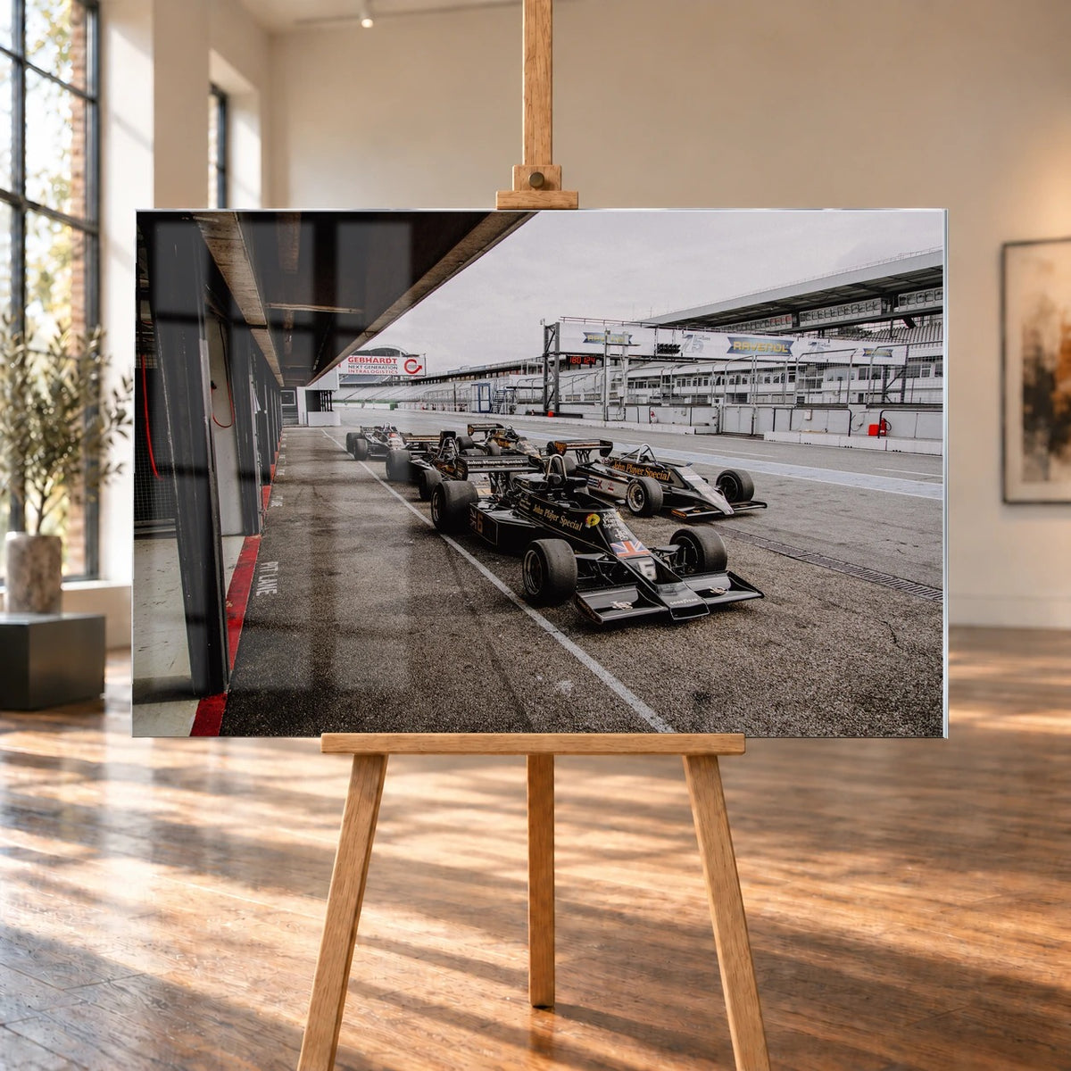 Mehrere historische Lotus-Rennwagen rollen in enger Formation an der Boxengasse entlang; der Blick von oben zeigt die Staffelung. Motiv „Trackside Moment“ als Acrylglas-Wandbild, auf einer Holzstaffelei in einem hellen Galerieraum.