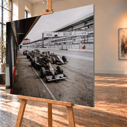 Mehrere historische Lotus-Rennwagen rollen in enger Formation an der Boxengasse entlang; der Blick von oben zeigt die Staffelung. Motiv „Trackside Moment“ als Alu gebürstet-Wandbild, schräg von der Seite auf einer Holzstaffelei.