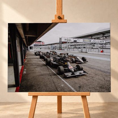 Mehrere historische Lotus-Rennwagen rollen in enger Formation an der Boxengasse entlang; der Blick von oben zeigt die Staffelung. Motiv „Trackside Moment“ als Leinwand-Wandbild, frontal auf einer Holzstaffelei.