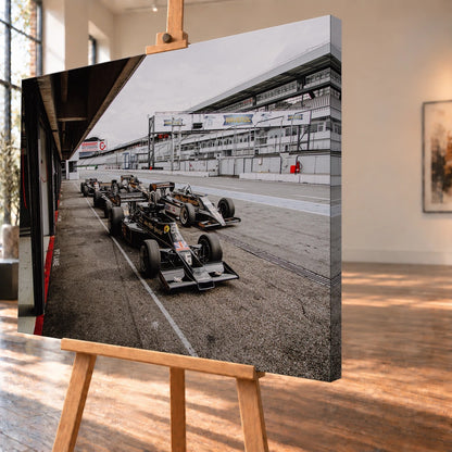 Mehrere historische Lotus-Rennwagen rollen in enger Formation an der Boxengasse entlang; der Blick von oben zeigt die Staffelung. Motiv „Trackside Moment“ als Leinwand-Wandbild, schräg von der Seite auf einer Holzstaffelei.